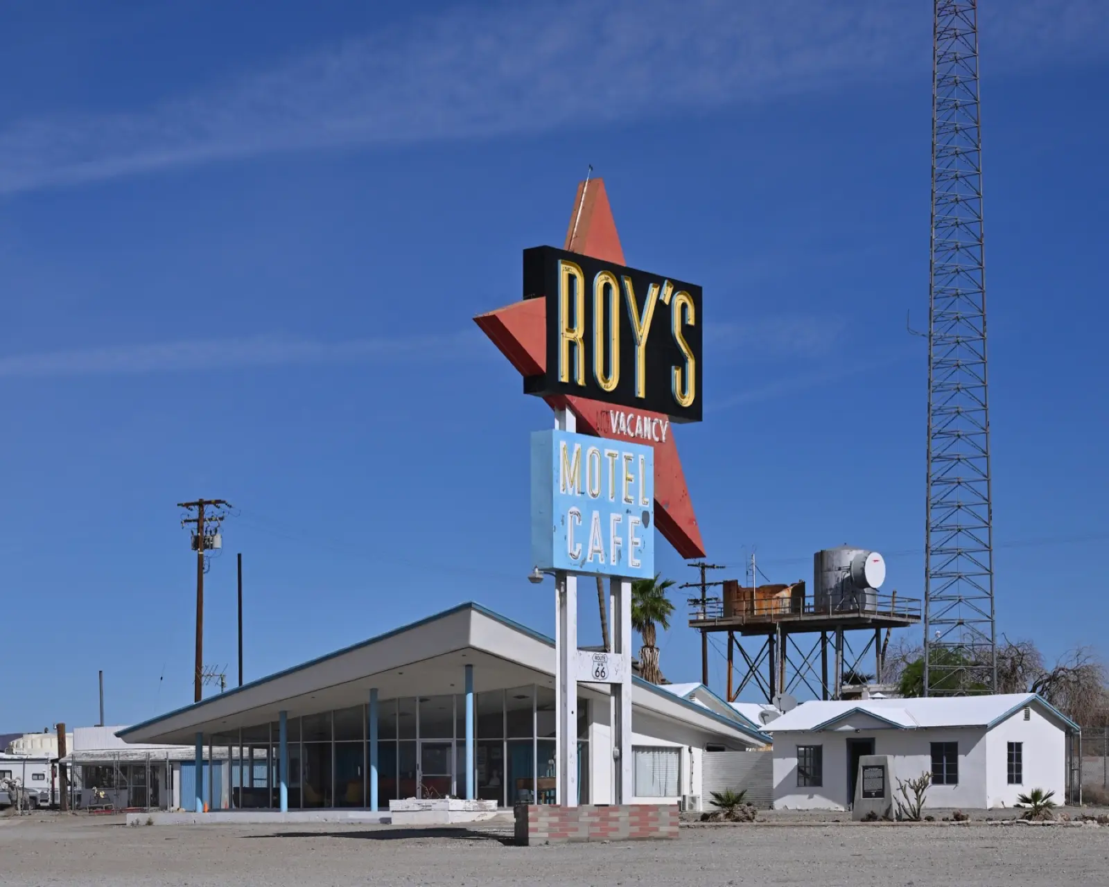 The tall Roy's Motel Cafe neon sign with a red boomerang arrow in front of the low glass-walled lobby building in Amboy, California, under a blue desert sky