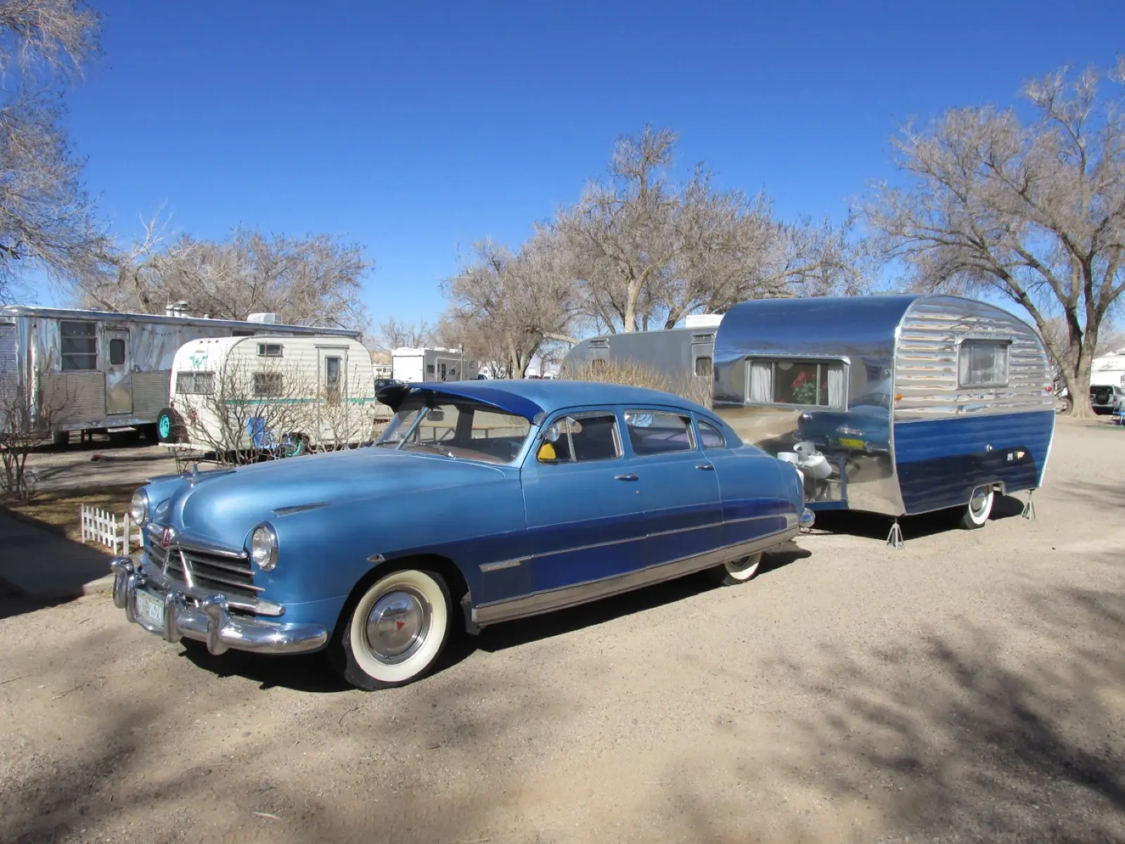 A blue 1950s Hudson Commodore towing a polished vintage camper trailer, with more vintage trailers parked behind, at Enchanted Trails in Albuquerque