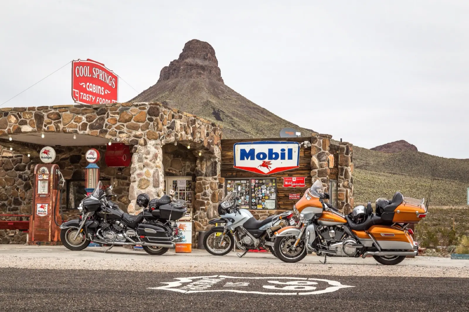 Three touring motorcycles parked in front of the stone Cool Springs station on Route 66 in Arizona, with a Route 66 shield painted on the pavement